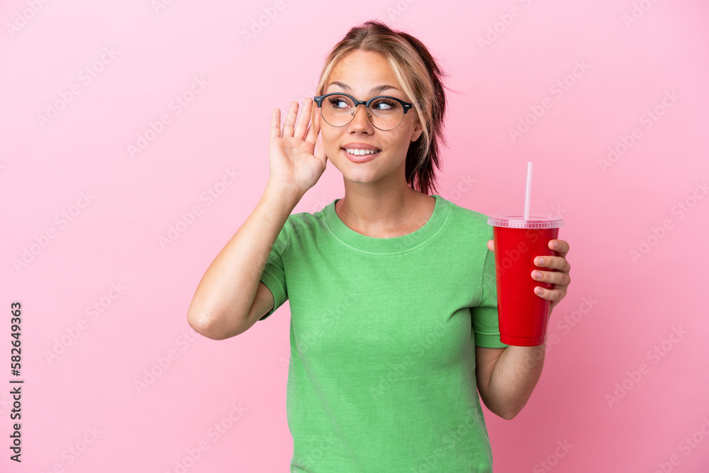 Young Russian woman holding a refreshment isolated on pink background listening to something by putting hand on the ear Young Russian woman holding a refreshment isolated on pink background listening to something by putting hand on the ear