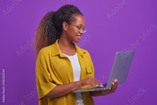 Young positive African American woman blogger with laptop in hand types article for own website and maintains online diary discussing popular topics with subscribers stands in lilac studio photo