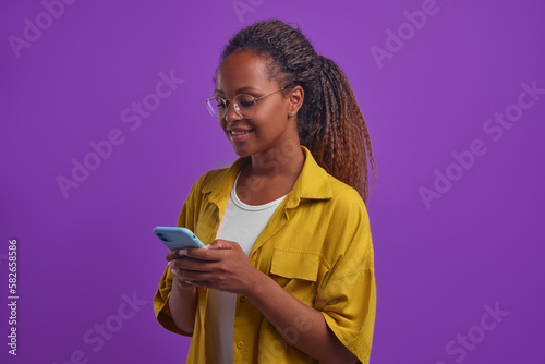Young attractive curly African American woman SMM specialist works with mobile phone as PR manager responding to user comments in social networks stands on lilac background. Marketing, freelance