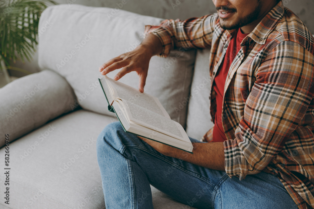 Cropped young Indian man wear casual clothes reading book novel prepare for exam sits on grey sofa couch stay at home hotel flat rest relax spend free spare time in living room indoor. Lounge concept.