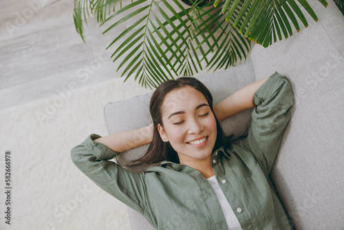 Top view young woman of Asian ethnicity wears casual clothes hold hands behind neck close eyes sits on grey sofa couch stay at home hotel flat rest relax spend free spare time in living room indoor. photo