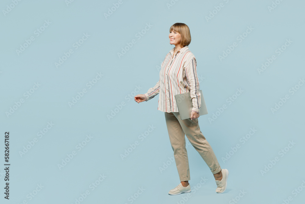 Full body side profile view elderly woman 50s years old wear shirt hold closed laptop pc computer look aside walk going strolling isolated on plain pastel blue cyan color background studio portrait Full body side profile view elderly woman 50s years old wear shirt hold closed laptop pc computer look aside walk going strolling isolated on plain pastel blue cyan color background studio portrait