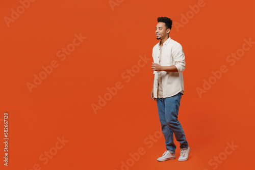 Full body smiling happy fun cheerful cool young man of African American ethnicity wearing light shirt casual clothes walking going isolated on orange red background studio portrait. Lifestyle concept.
