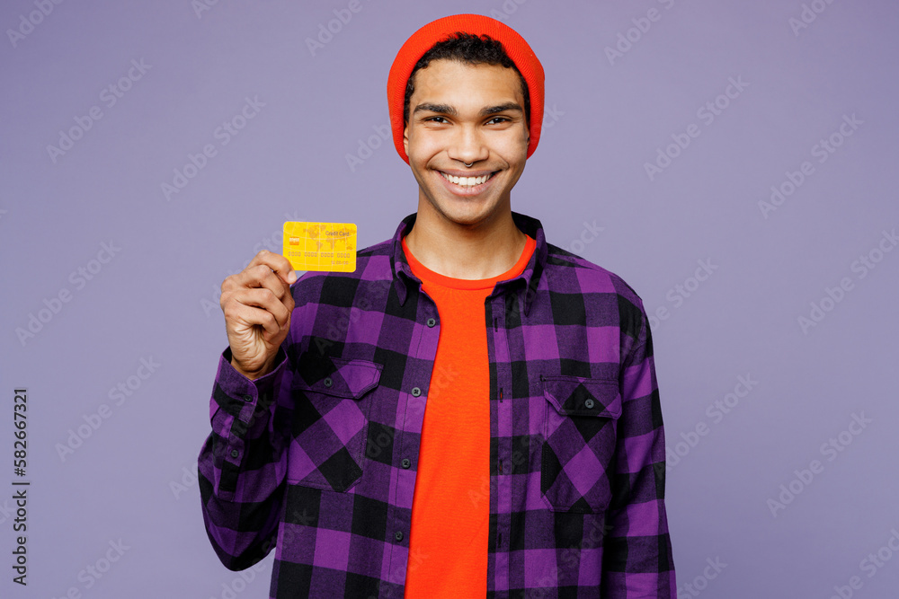 Young smiling happy fun man of African American ethnicity wear casual shirt orange hat hold in hand mock up of credit bank card isolated on plain pastel light purple color background studio portrait. Young smiling happy fun man of African American ethnicity wear casual shirt orange hat hold in hand mock up of credit bank card isolated on plain pastel light purple color background studio portrait.
