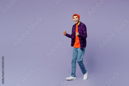 Full body young man of African American ethnicity wear casual shirt orange hat doing winner gesture celebrate clenching fists say yes isolated on plain pastel purple color background studio portrait.