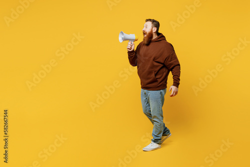Full body young redhead caucasian man wearing brown hoody casual clothes hold in hand megaphone scream announces discounts sale Hurry up isolated on plain yellow background studio. Lifestyle concept.