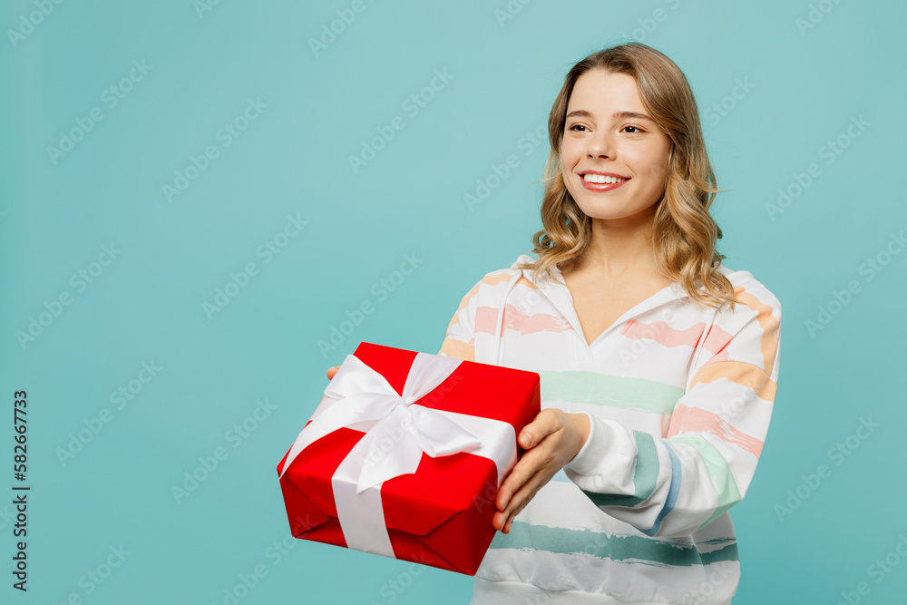 Young smiling happy cheerful blonde woman wear striped hoody hold in hand give present box with gift ribbon bow isolated on plain pastel light blue cyan background studio portrait. Lifestyle concept. Young smiling happy cheerful blonde woman wear striped hoody hold in hand give present box with gift ribbon bow isolated on plain pastel light blue cyan background studio portrait. Lifestyle concept.