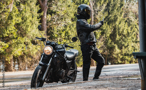 man in a leather motorcycle jacket with a motorcycle custom cafe racer in a helmet on a forest road with a smartphone.
