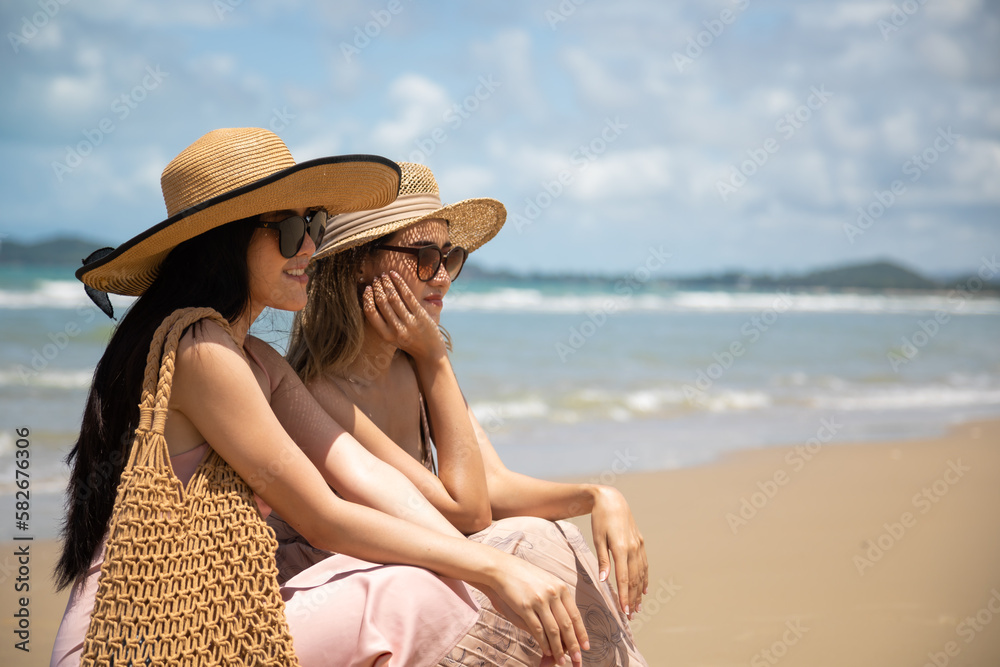Two happy young asian women sit on logs by the sea. Vacation trip summer holiday.