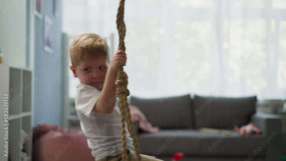Calm toddler boy rides on swing in light living room with sofa and bean bag chair. Thoughtful blond boy holds on tight to ropes resting at home Calm toddler boy rides on swing in light living room with sofa and bean bag chair. Thoughtful blond boy holds on tight to ropes resting at home