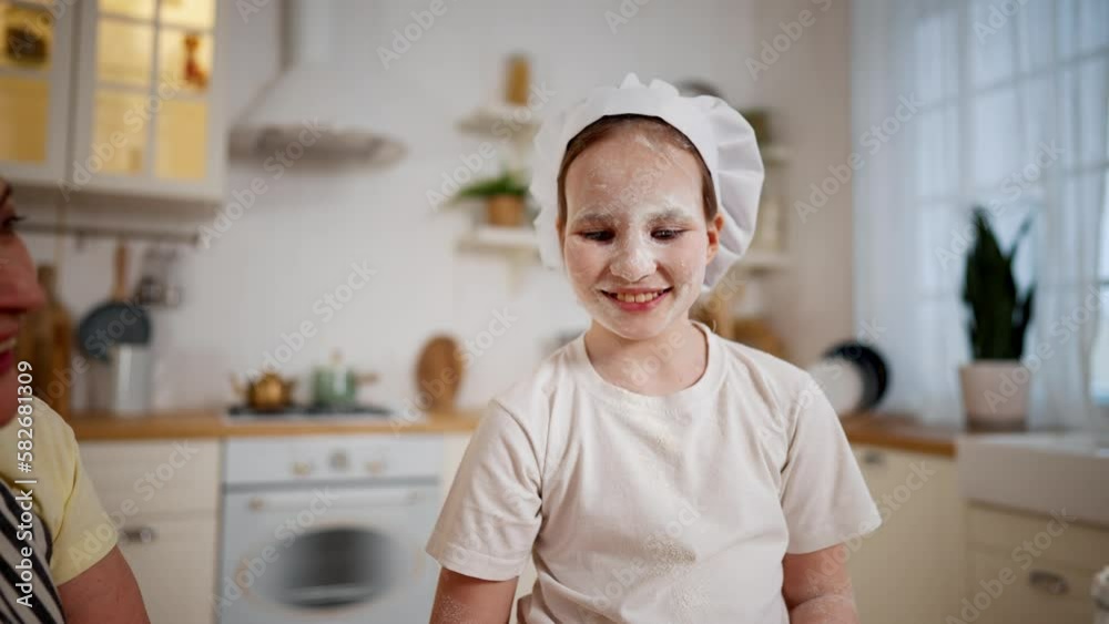 Smiling happy child girl helps cook to granny in kitchen. Her face dirty in flour, granny touches her nose. Cooking together, family sweet moments. Portrait of pre-teen laughing girl in cook's cap Smiling happy child girl helps cook to granny in kitchen. Her face dirty in flour, granny touches her nose. Cooking together, family sweet moments. Portrait of pre-teen laughing girl in cook's cap