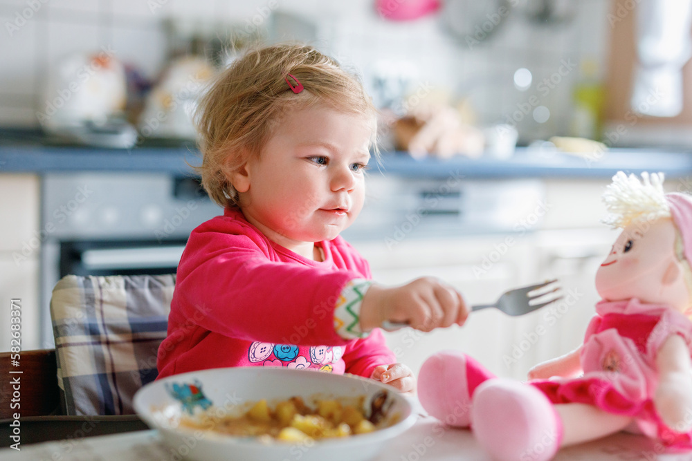 Adorable baby girl eating from fork vegetables and pasta. food, child, feeding and development concept. Cute toddler, daughter with spoon sitting in highchair and learning to eat by itself.