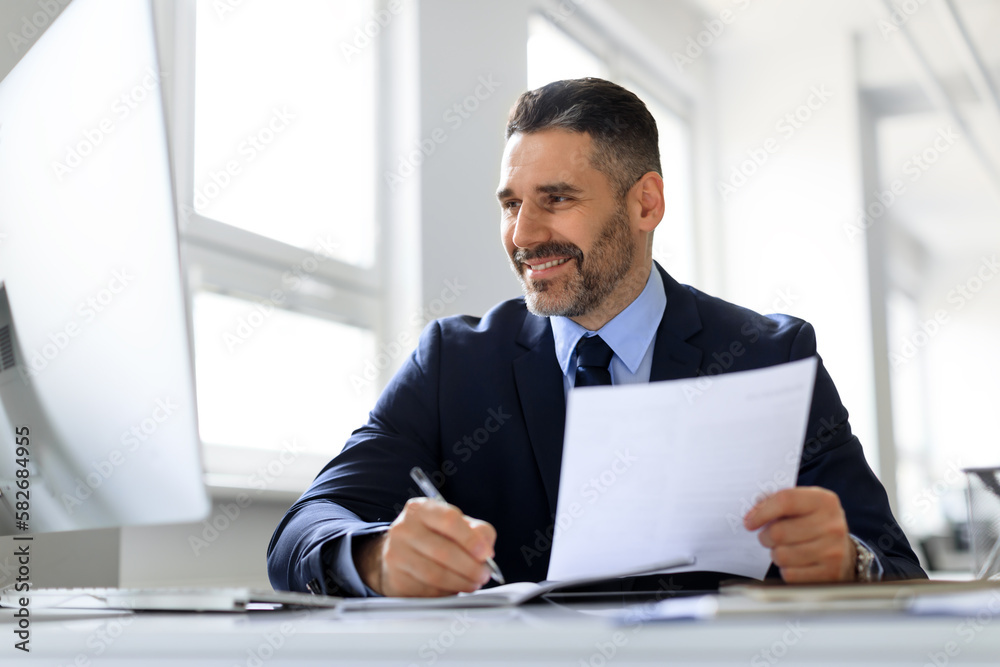 Happy middle aged businessman working with computer and documents in office, male entrepreneur in suit sitting at desk Happy middle aged businessman working with computer and documents in office, male entrepreneur in suit sitting at desk