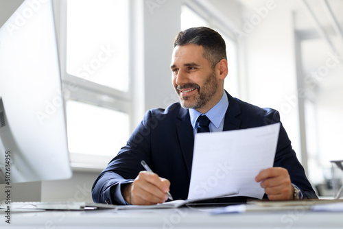 Happy middle aged businessman working with computer and documents in office, male entrepreneur in suit sitting at desk