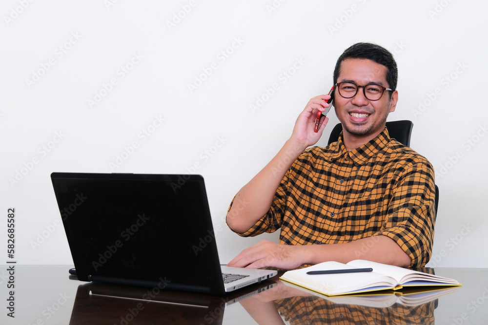 Asian man sitting in his working desk showing happy face while talking on phone Asian man sitting in his working desk showing happy face while talking on phone