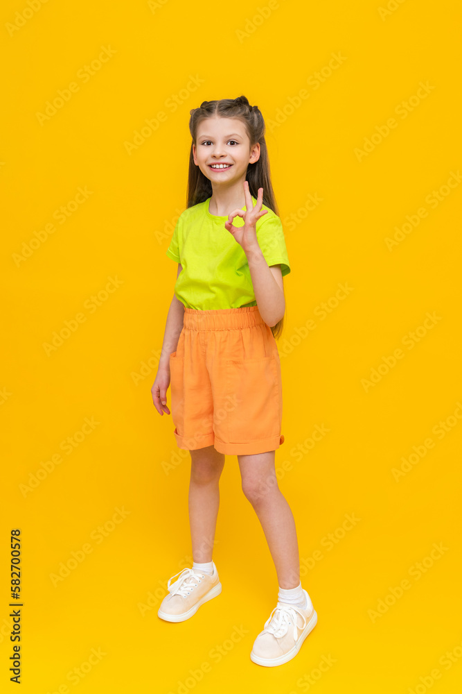 A child in shorts and a T-shirt. Happy childhood. A little girl in full height with long hair rejoices in the summer warmth inside and points to your advertisement on a yellow isolated background. A child in shorts and a T-shirt. Happy childhood. A little girl in full height with long hair rejoices in the summer warmth inside and points to your advertisement on a yellow isolated background.