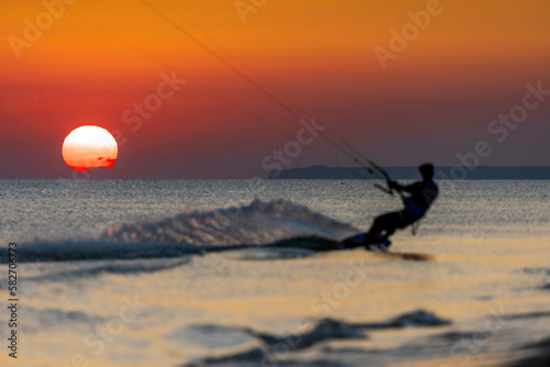 Scenic sunset seascape with silhouette of kitesurfer on foreground splashing by shoreline. Sport, summer activities, travel, adventure and exploration concept.