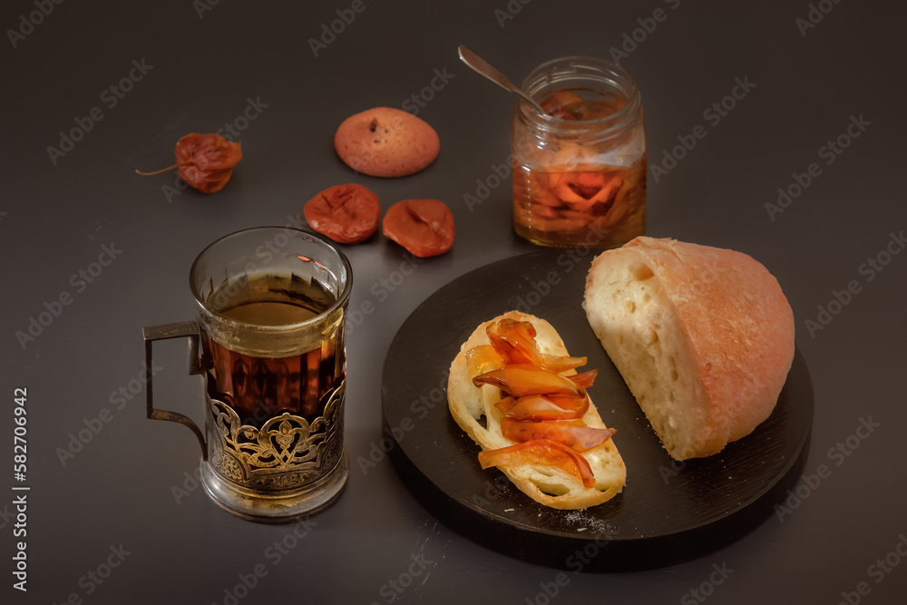 Rustic breakfast with a piece of bread and jam, a glass of hot tea in an antique silver cup holder and a jar of jam on a gray wooden table