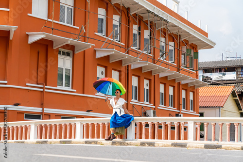 Asian guy drag queen in woman clothes, makeup and sunglasses holding rainbow umbrella walking down city street. Diversity sexual equality, lgbtq pride people and transgender cross-dressing concept.