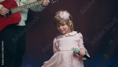 Little girl on stage in vintage dress, she sings into microphone and dances, her father plays acoustic guitar. Color music is shining. Performance on stage. Early development of children