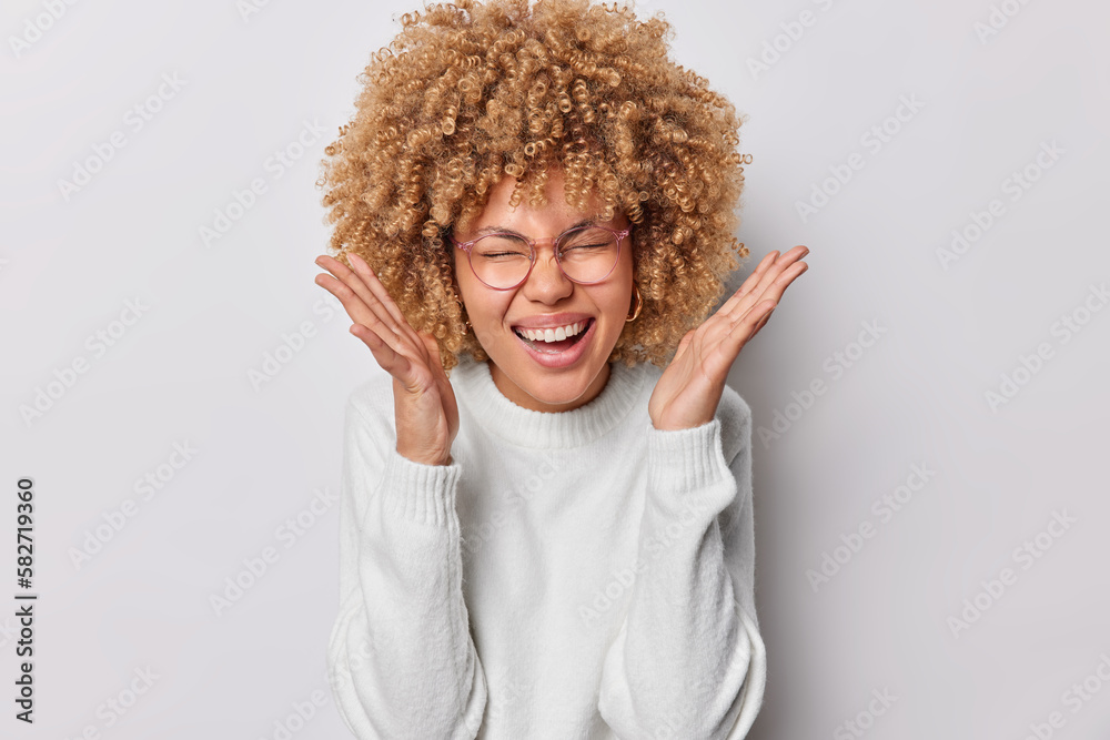 Emotional curly haired woman exclaims loudly keeps palms raised near face smiles broadly shows perfect teeth reacts to something awesome dressed in casual jumper isolated over white background Emotional curly haired woman exclaims loudly keeps palms raised near face smiles broadly shows perfect teeth reacts to something awesome dressed in casual jumper isolated over white background