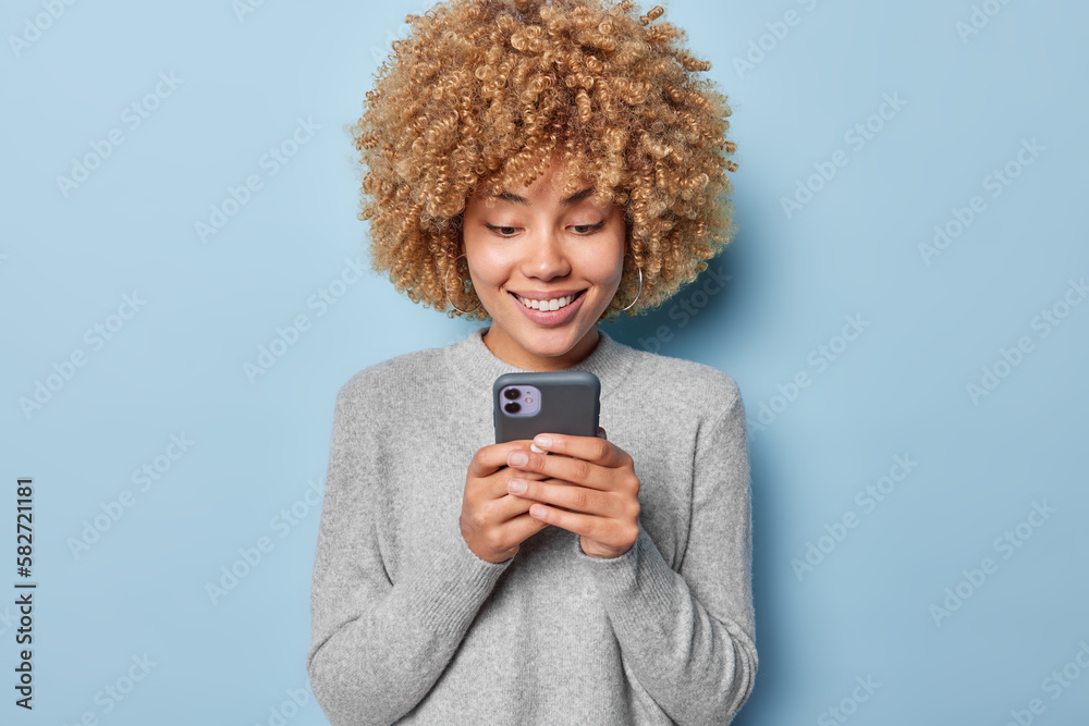 Horizontal shot of pretty curly haired woman uses mobile phone uses social media app chats on cellular smiles positively dressed in casual grey jumper isolated over blue background. Modern technology