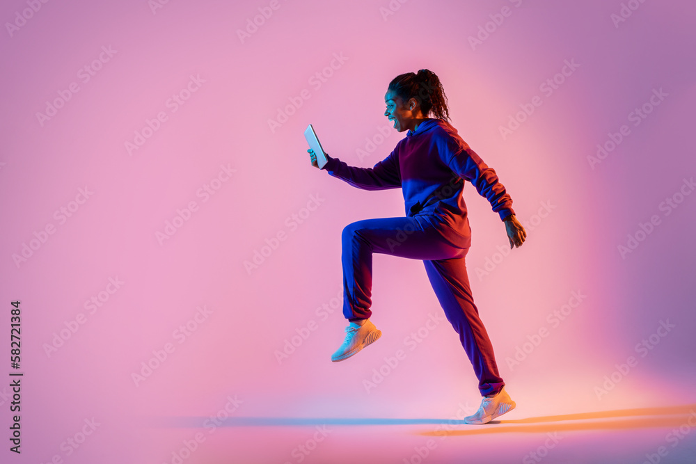 Full body length shot of black lady jumping or running, using cellphone over pink neon background, side view, copy space