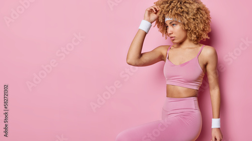 Horizontal shot of motivated curly haired sportswoman wears top and leggings headband wristband looks seriously aside prepares for fitness training isolated over pink background blank space for promo photo