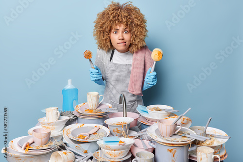 Фотография Worried busy housewife bites lips and looks at camera holds cleaning brush wears