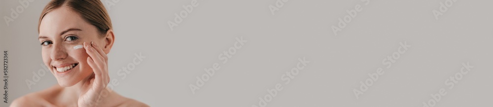 Beautiful young redhead woman applying cream on face and smiling against grey background