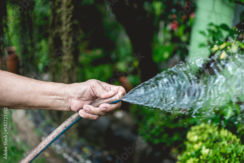 Hand of an old lady holding a rubber hose and blocking the tip to get a spray of water. Outdoor or garden setting.