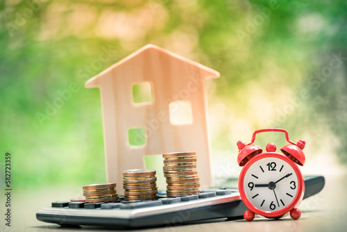 Stacked gold coins with growing interest put on the calculator and  and clock with time and blur of home model on sunlight in the public park, Business investment and loans for real estate concept.