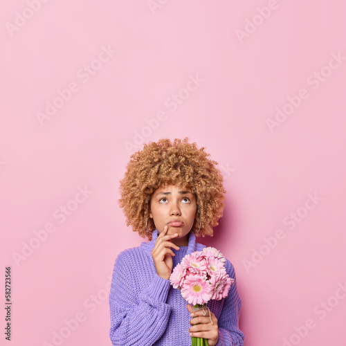 Vertical shot of pensive curly haired woman purses lips focused above with displeased expression holds gerbera flowers dressed in knitted jumper isolated over pink background copy space overhead