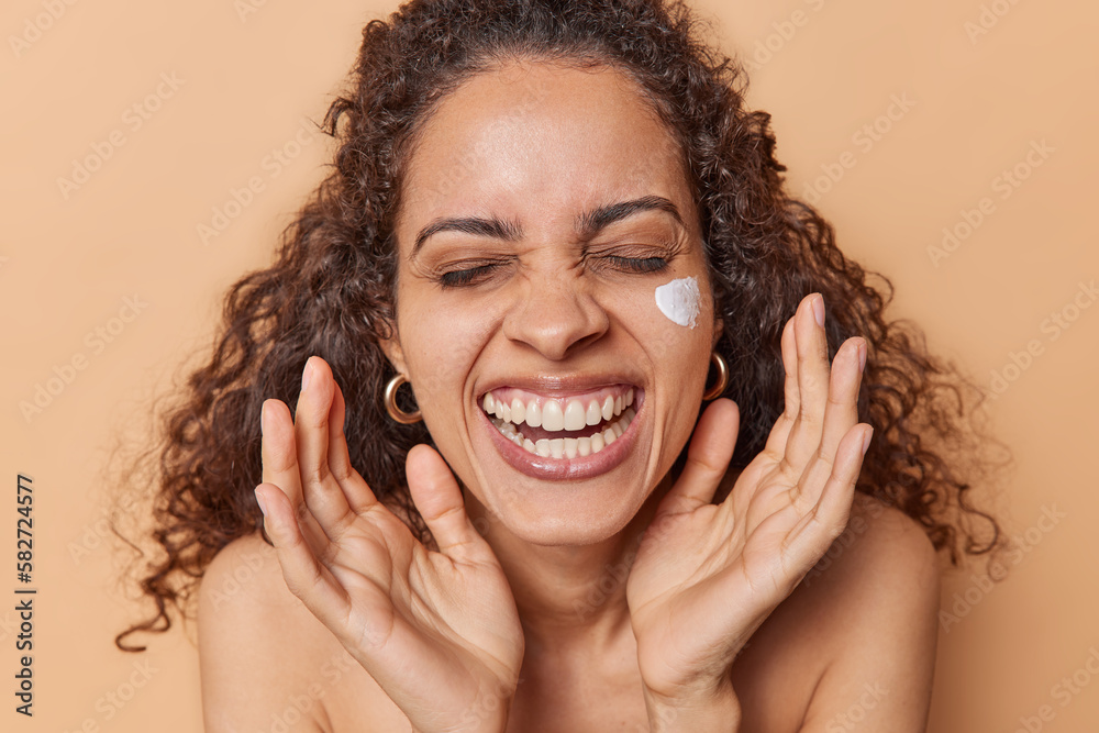 Fototapeta premium Daily skin care concept. Overjoyed curly haired woman applies facial cream for moisturising takes care of skin smiles toothily stands half naked isolated over brown background uses beauty product