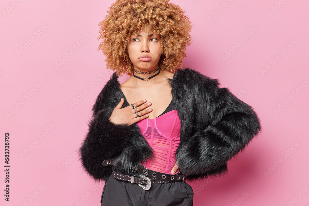 Horizontal shot of displeased curly haired young woman looks unhappily aside dressed in fur coat an black trousers with belt purses lips isolated over pink background feels tired of everything Horizontal shot of displeased curly haired young woman looks unhappily aside dressed in fur coat an black trousers with belt purses lips isolated over pink background feels tired of everything