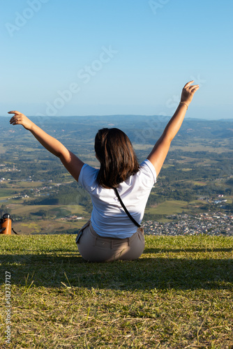 Mulher sentada de costas olhando para uma paisagem de cima do morro, sentada na grama verde e leventando os braços em forma de V photo