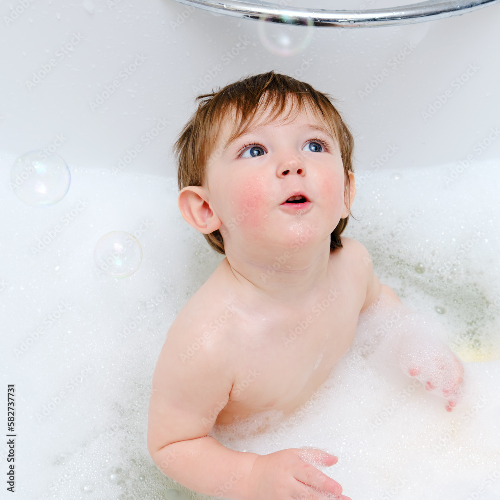 A surprised child is playing with soap bubbles while sitting in a white home bathtub. Curious toddler baby boy bathes in the bathroom. Kid aged one year six months A surprised child is playing with soap bubbles while sitting in a white home bathtub. Curious toddler baby boy bathes in the bathroom. Kid aged one year six months