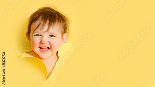 Happy baby in a hole on a paper yellow background. Torn child's head studio background, copy space. Kid aged one year six months photo