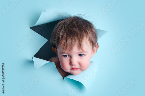 Sad baby in a hole on a paper blue background. Torn child's head studio background, copy space. Kid aged one year six months photo