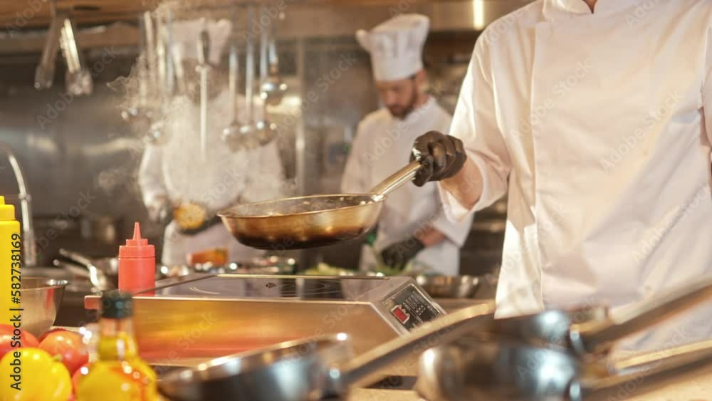 Close-up portrait of delicious food being frying on pan and mixed by handsome young caucasian professional chef in white uniform. Attractive cook cooking tasty dishes in modern kitchen.