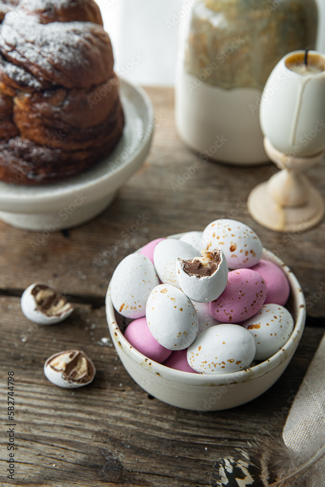 Bowl with white, pink and foiled chocolate eggs, homemade Easter sweet bread, pink tulips and feathers on rustic wooden table.