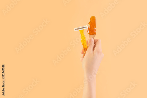 Female hand holding spatula with sugaring paste and razor on beige background