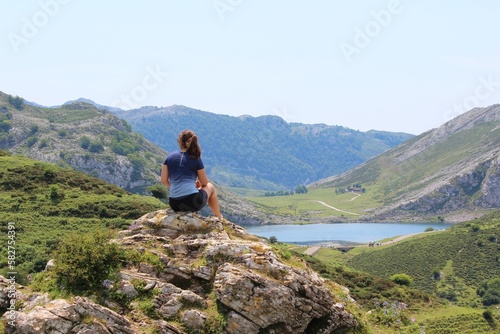 Young woman sitting on a rock after hiking and doing sports. In the background, big mountains and a lake. Beautiful green fields are seen and she is wearing a blue t-shirt.