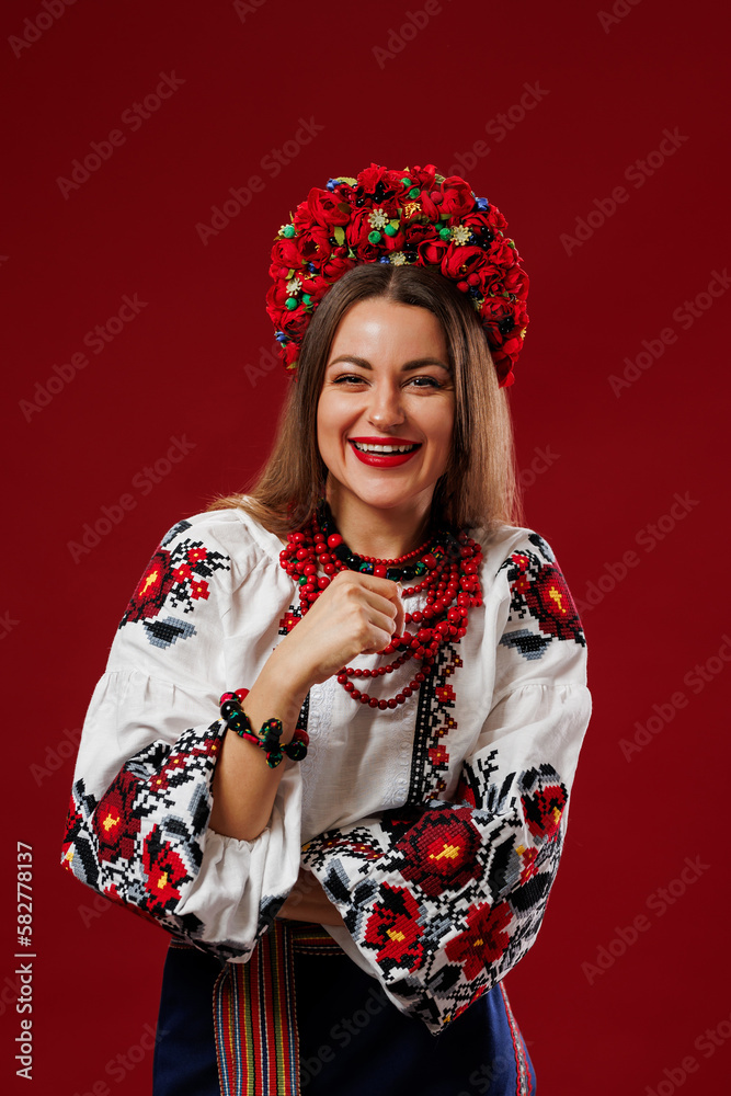 Fototapeta premium Portrait of ukrainian woman in traditional ethnic clothing and floral red wreath on viva magenta studio background. Ukrainian national embroidered dress call vyshyvanka. Pray for Ukraine