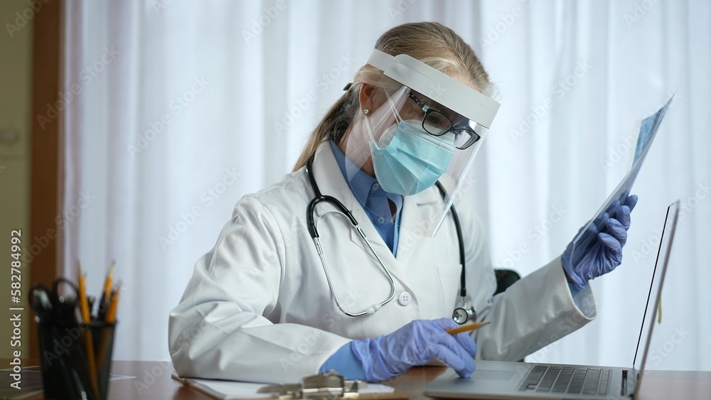Female doctor wearing face shield face mask and surgical gloves looks at X-ray image of patient. Woman physician wearing PPE personal healthcare in office. Female doctor wearing face shield face mask and surgical gloves looks at X-ray image of patient. Woman physician wearing PPE personal healthcare in office.