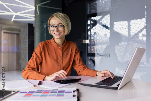 Portrait of successful financier woman, businesswoman behind paper accounting work smiling and looking at camera, female worker happy at workplace using laptop inside office, blonde in glasses.