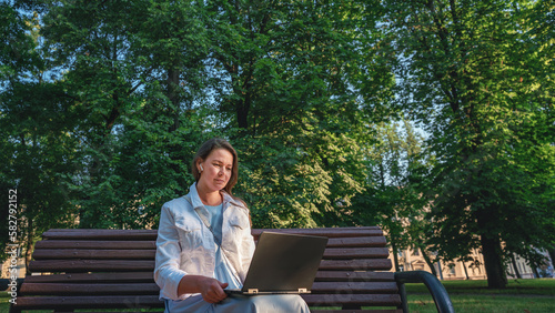 Adult woman seated on wooden bench in urban park is working by laptop. Freelance and distant job concept.