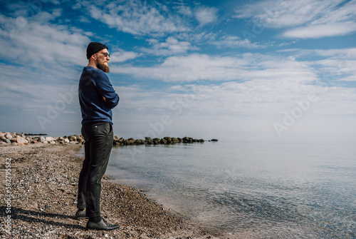 Fototapeta Naklejka Na Ścianę i Meble -  cooler junger Mann mit Bart, Sonnenbrille und Mütze steht bei blauem Himmel am Strand und sieht in die Ferne.