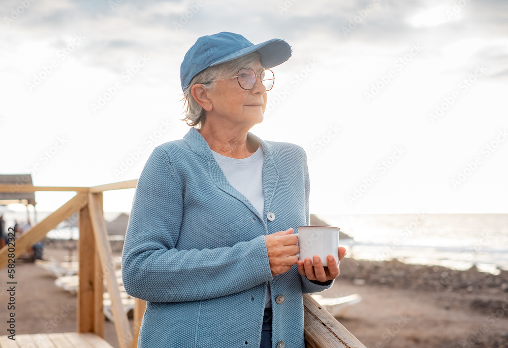 Portrait of pensive attractive senior woman in sea vacation standing at beach at sunset enjoying a cup of tea