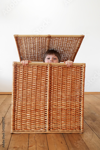 Smile, happy little boy sit in brown wicker basket and peeps out. Cute kid play and hide in basket. Development and education of child at home